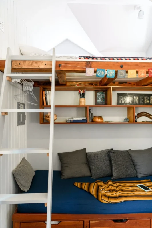Loft bed with white ladder and built-in wooden shelving cubbies in a tiny apartment